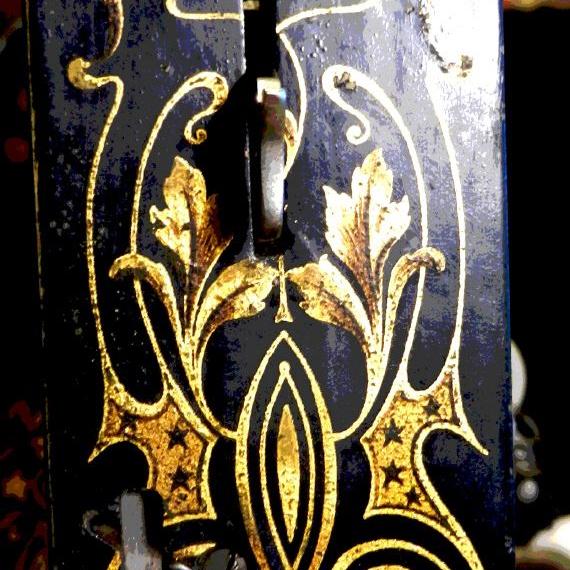 Ein dunkler, kunstvoll verzierter Nähmaschinenfuss mit goldfarbenen ornamentalen Verzierungen und sichtbaren mechanischen Komponenten.
