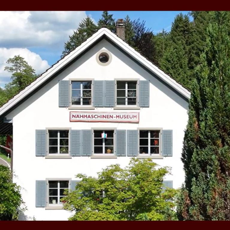 Ein traditionelles, weißes Museumshaus mit dem Schild Nahmaschinen-Museum ist in eine natürliche, bewaldete Landschaft eingebettet.