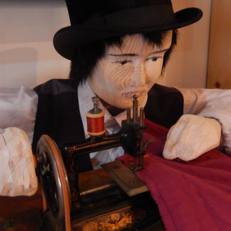 Eine lebensgroße Puppe mit Hut, die an einer alten Nähmaschine sitzt und zu nähen scheint.